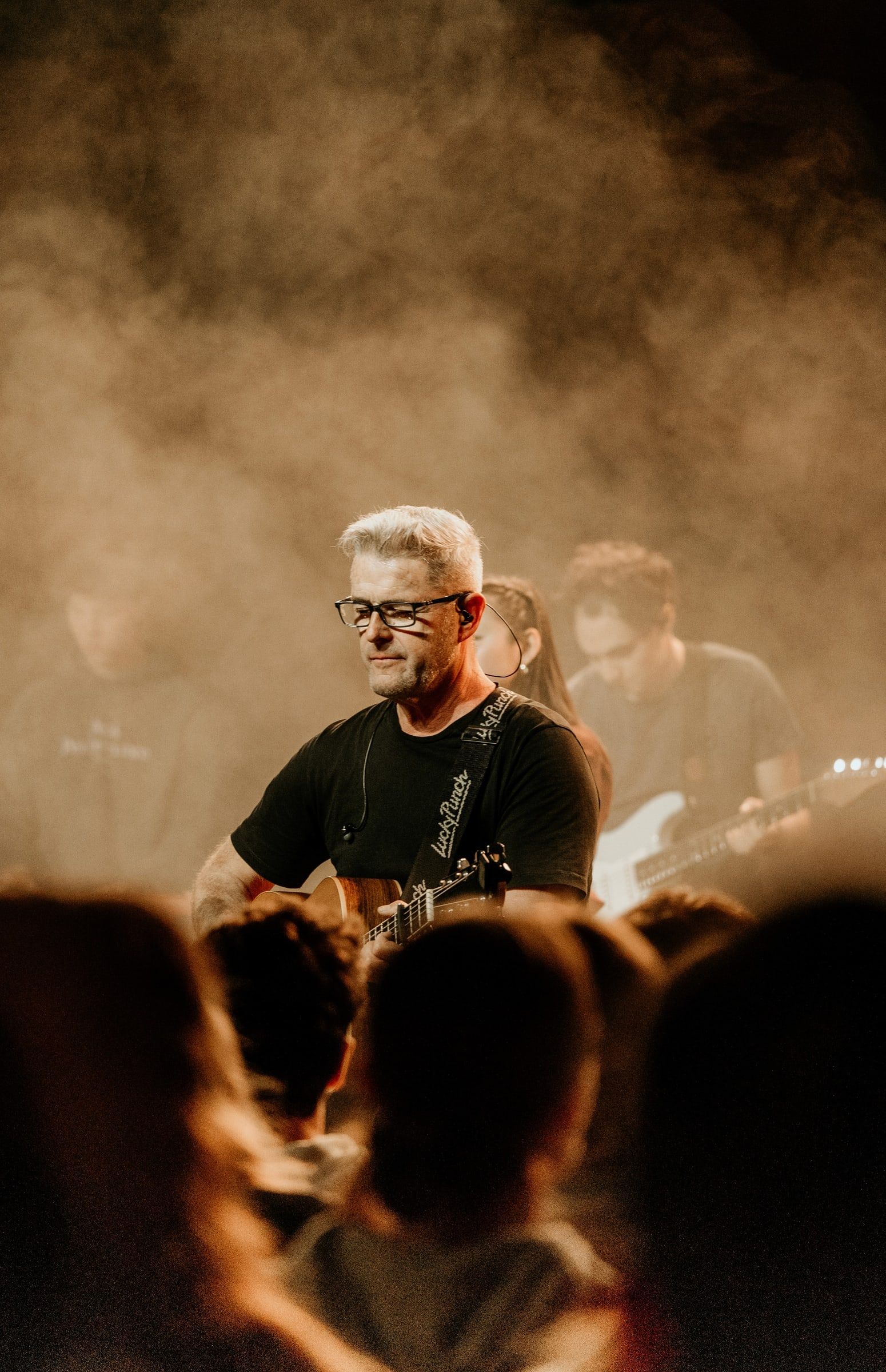 Gitarrist mit Brille spielt im warmen Bühnenlicht, Publikum im Vordergrund
