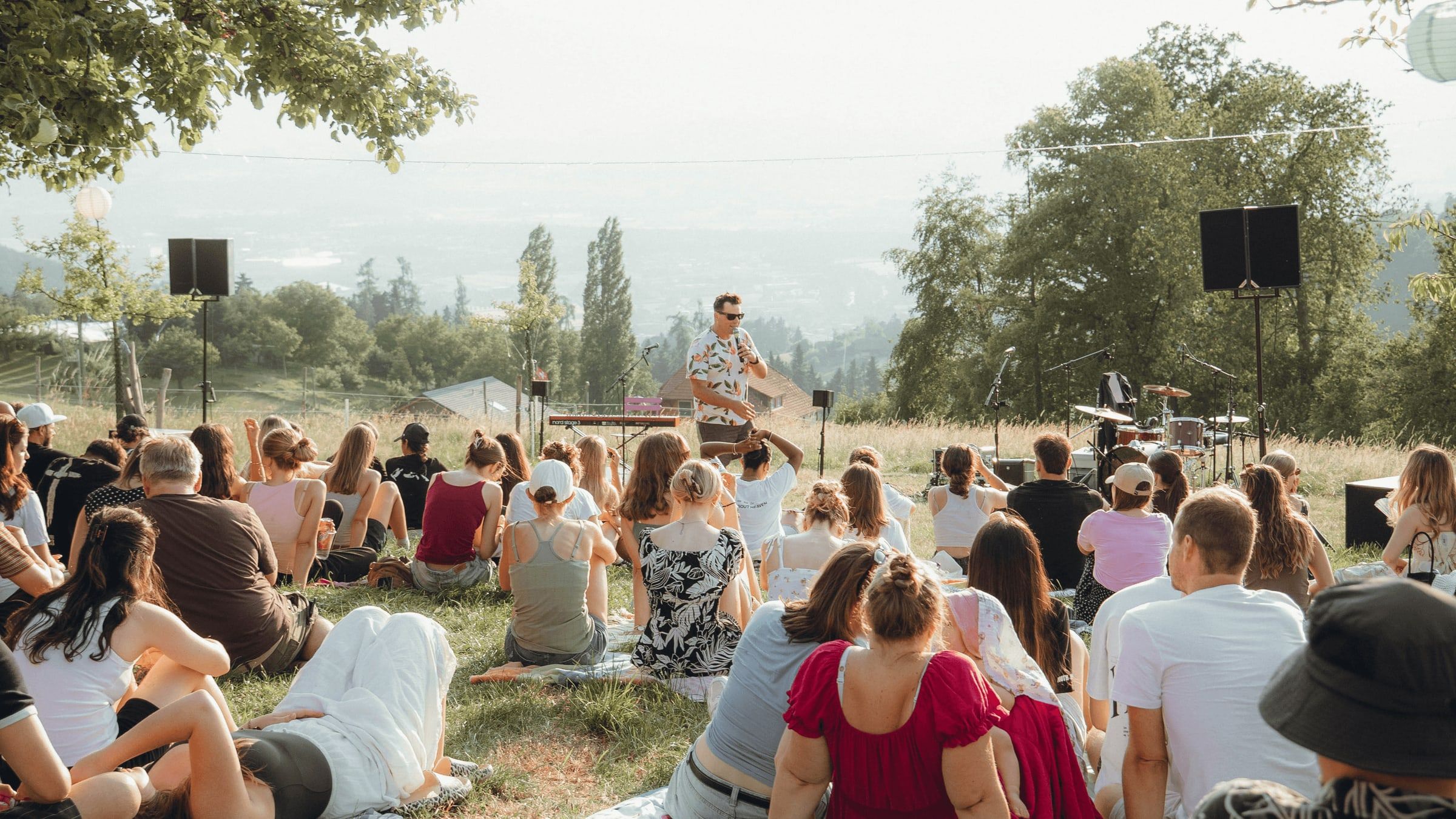 Publikum sitzt auf einer Wiese vor einer kleinen Open-Air-Bühne